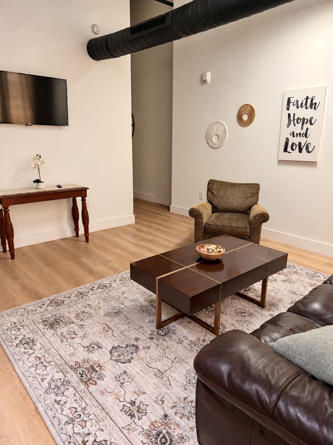 A modern living room with a flat-screen TV, a wooden coffee table, a brown armchair, a patterned rug, and a leather sofa.