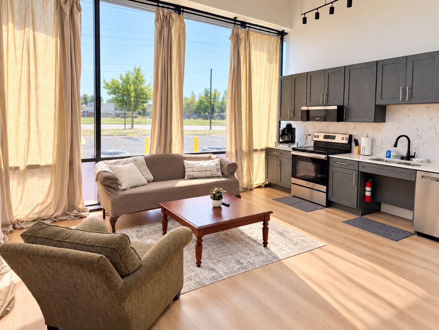 A modern living room with a sofa, armchair, and coffee table, a kitchen with dark cabinets and appliances, and large windows with curtains.