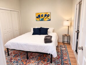 Un dormitorio con cama, almohadas, manta, alfombra, un cuadro, una lámpara y una puerta.