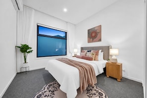 A modern bedroom with a large window, a bed with multiple pillows, a bedside table with a lamp, and a decorative plant. - 2 New Bayswater Homes | 8BR/6BA, Sleeps 16 (Bayswater)