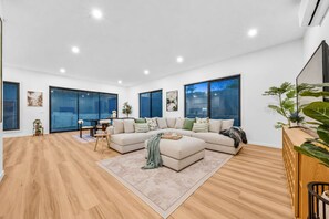 A spacious living room with a large sectional sofa, a dining table, and a view of the outdoors through large windows. - 2 New Bayswater Homes | 8BR/6BA, Sleeps 16 (Bayswater)