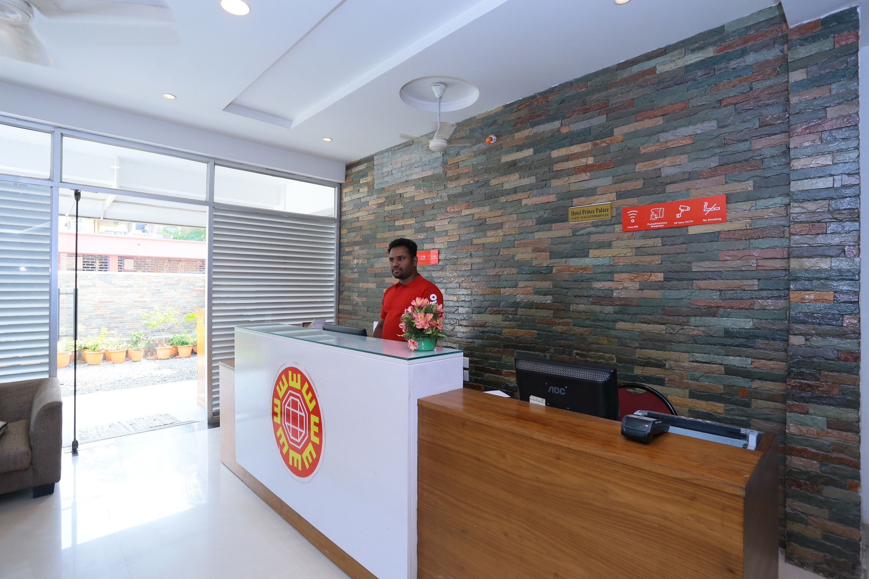 Una recepción de hotel con diseño moderno, un logotipo visible y un cartel que indica servicios como Wi-Fi gratuito, desayuno y cámaras de seguridad disponibles las 24 horas.