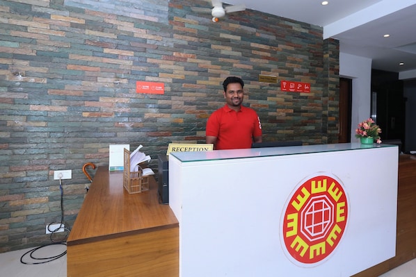 A reception desk with a stone wall backdrop, a sign indicating check-in and check-out times, and a logo on the desk. - Super Townhouse Prince Palace (Trivandrum)