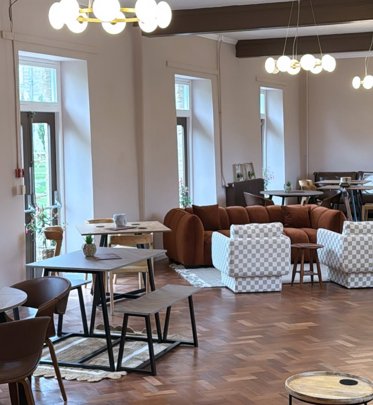 Una habitación amplia con suelo de madera en espiga, un sofá de cuero marrón y un sillón a cuadros. Hay mesas y sillas dispuestas para comer, y un ventanal deja entrar luz natural.