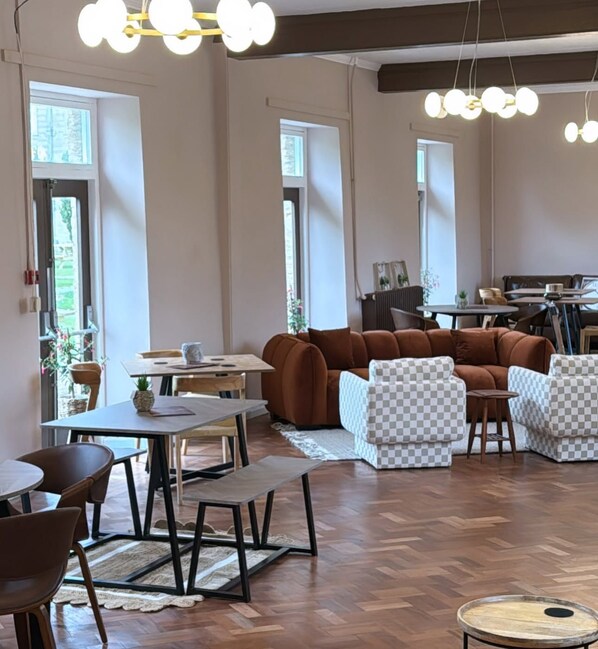 A spacious room with a wooden herringbone floor, a brown leather sofa, and a checkered armchair. There are tables and chairs arranged for dining, and a large window allows natural light to enter.