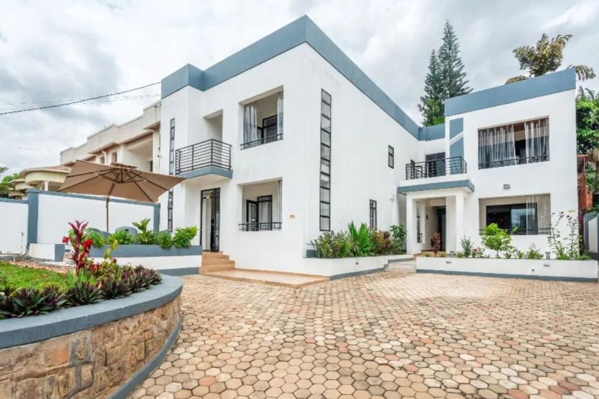 A modern two-story house with a paved courtyard, a balcony, and a garden with various plants.