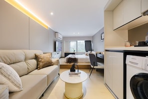 A modern hotel room with a sofa, a round coffee table, a bed with a headboard, a desk with a chair, a window with curtains, and a kitchen area with a dishwasher. - man  feng (Guangzhou)