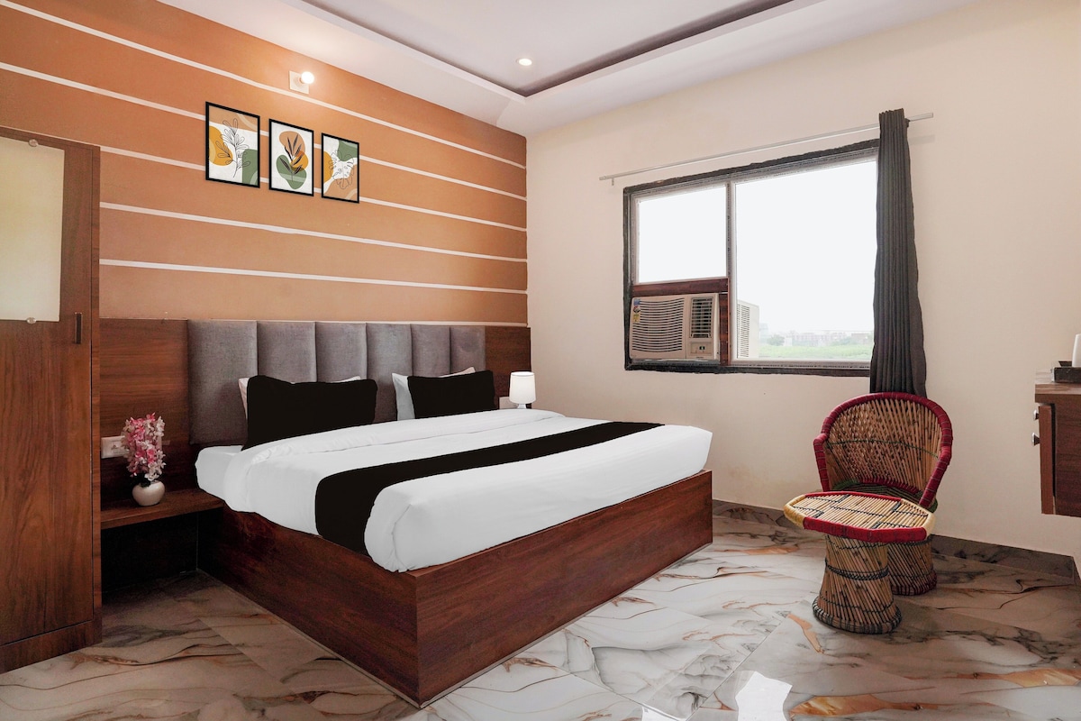 A hotel room with a wooden bed, a bedside table with a vase, a window with curtains, and a wicker chair.