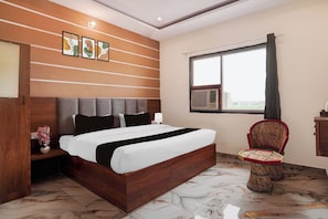 A hotel room with a wooden bed, a bedside table with a vase, a window with curtains, and a wicker chair. - Collection O Radha Krishna Vrindavan (Vrindavan)