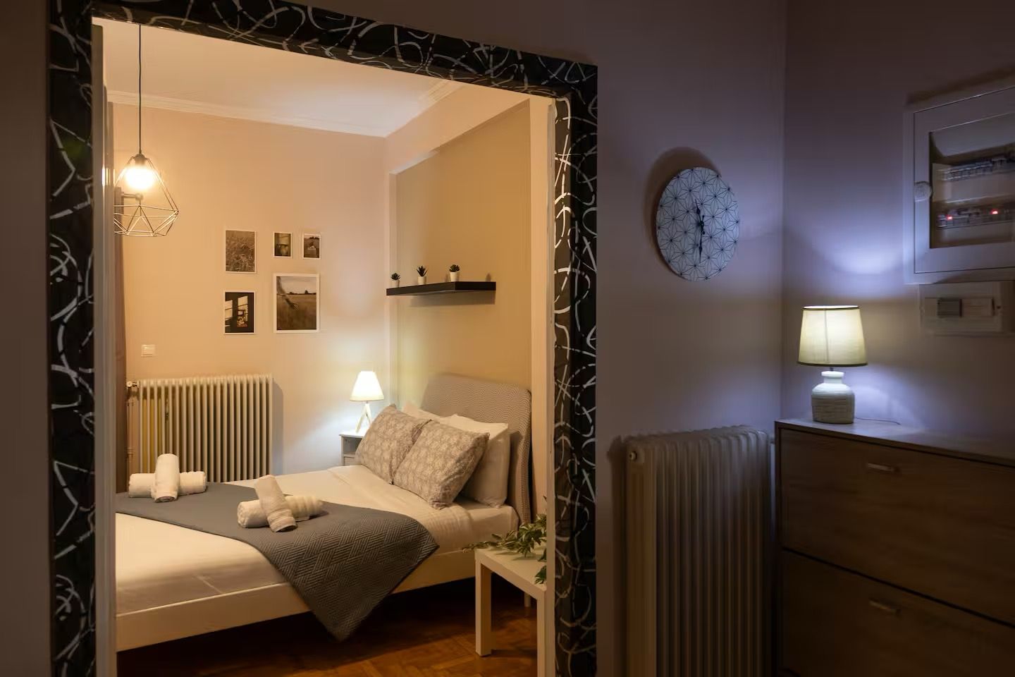 Ein Schlafzimmer mit einem Bett, Nachttischlampen, einem Heizkörper, einer Wanduhr und einem Regal mit kleinen Pflanzen.