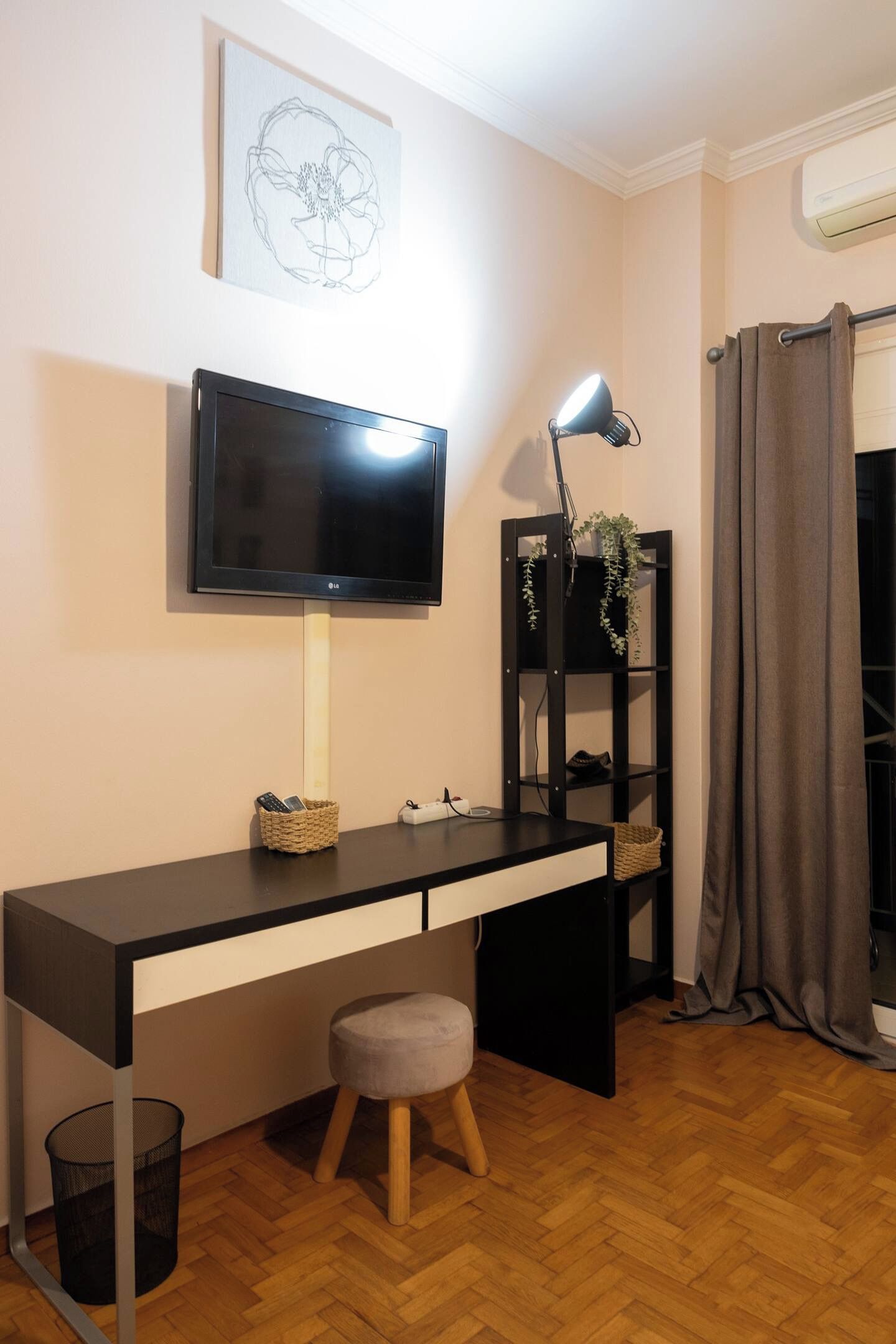 Ein modernes Zimmer mit einem wandmontierten Flachbildfernseher, einem Schreibtisch mit Lampe, einem kleinen Hocker und einem Papierkorb.
