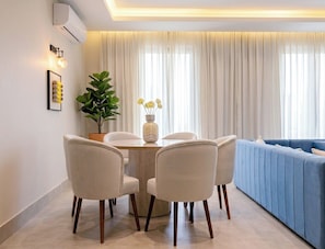 A modern living room with a blue sofa, a dining table with chairs, a potted plant, and a large window with curtains. - Wonderful 3-bedroom apartment with AC in vibrant riyadh (الرياض)