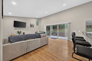 A modern living room with a sectional sofa, a flat-screen TV, a dining area, and a sliding glass door leading to a garden. - Escape to Pandapas Retreat! Enjoy a private hot tub, cozy comforts, and peaceful woodland scenery. (Newport)