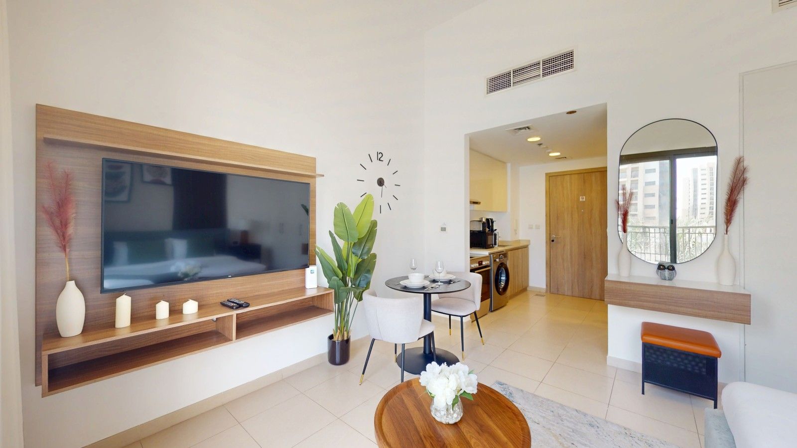 Un salon moderne avec un téléviseur à écran plat, un coin repas avec une table et des chaises, une cuisine en arrière-plan et un grand miroir au-dessus d’un banc.
