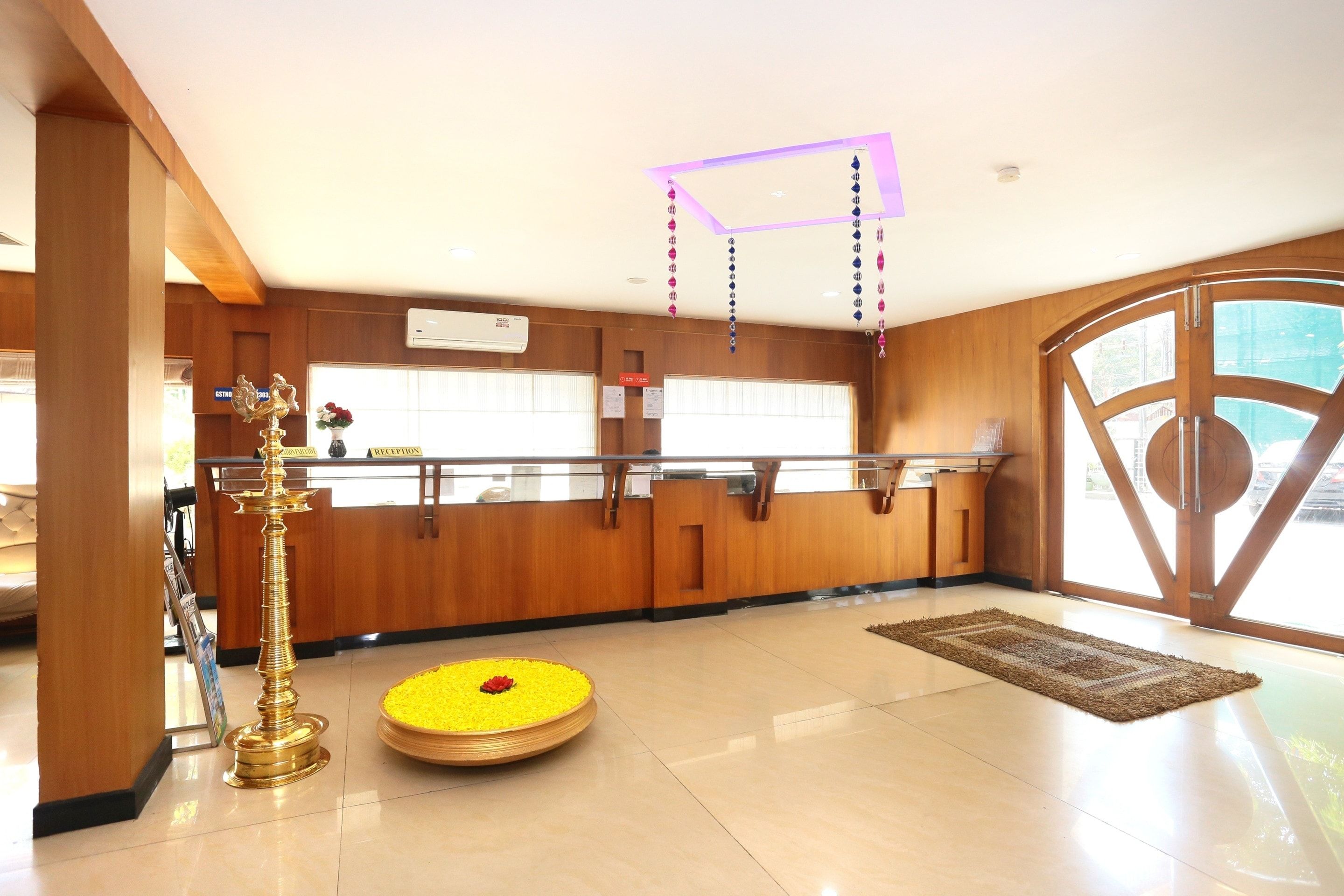 Un área de recepción con mostrador, una lámpara dorada decorativa, un pequeño arreglo floral amarillo y un ventanal con vistas al exterior.