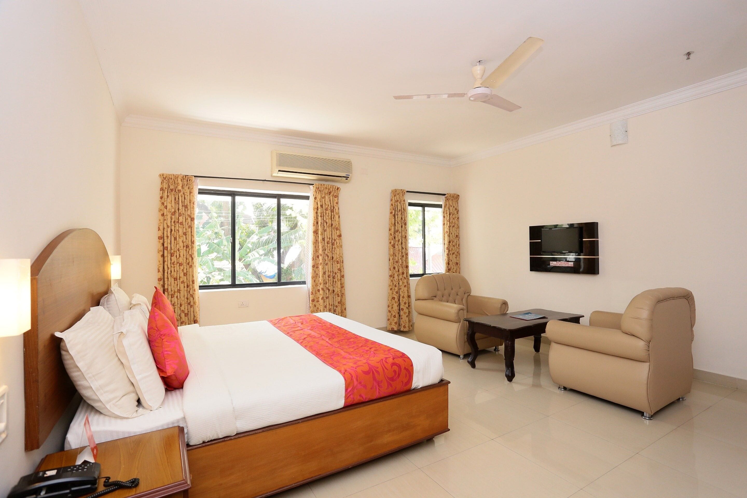 A hotel room with a bed, two armchairs, a sofa, a TV, and a ceiling fan.