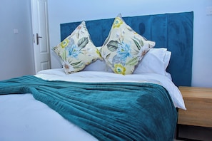 Uma cama com um cobertor azul-turquesa, duas almofadas florais e uma mesa de cabeceira de madeira.