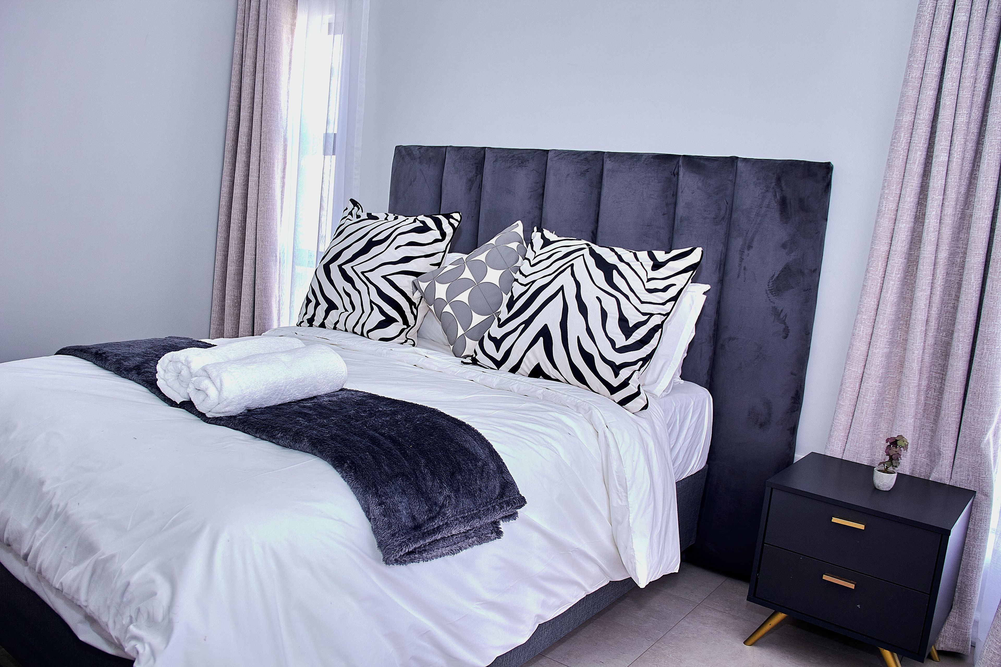 A modern bedroom with a large bed, a dark headboard, and a bedside table with drawers.