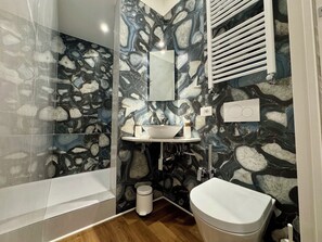 A bathroom with a stone-patterned wall, a white sink, a mirror, a toilet, and a shower area. - Vrbo Property (Roma)