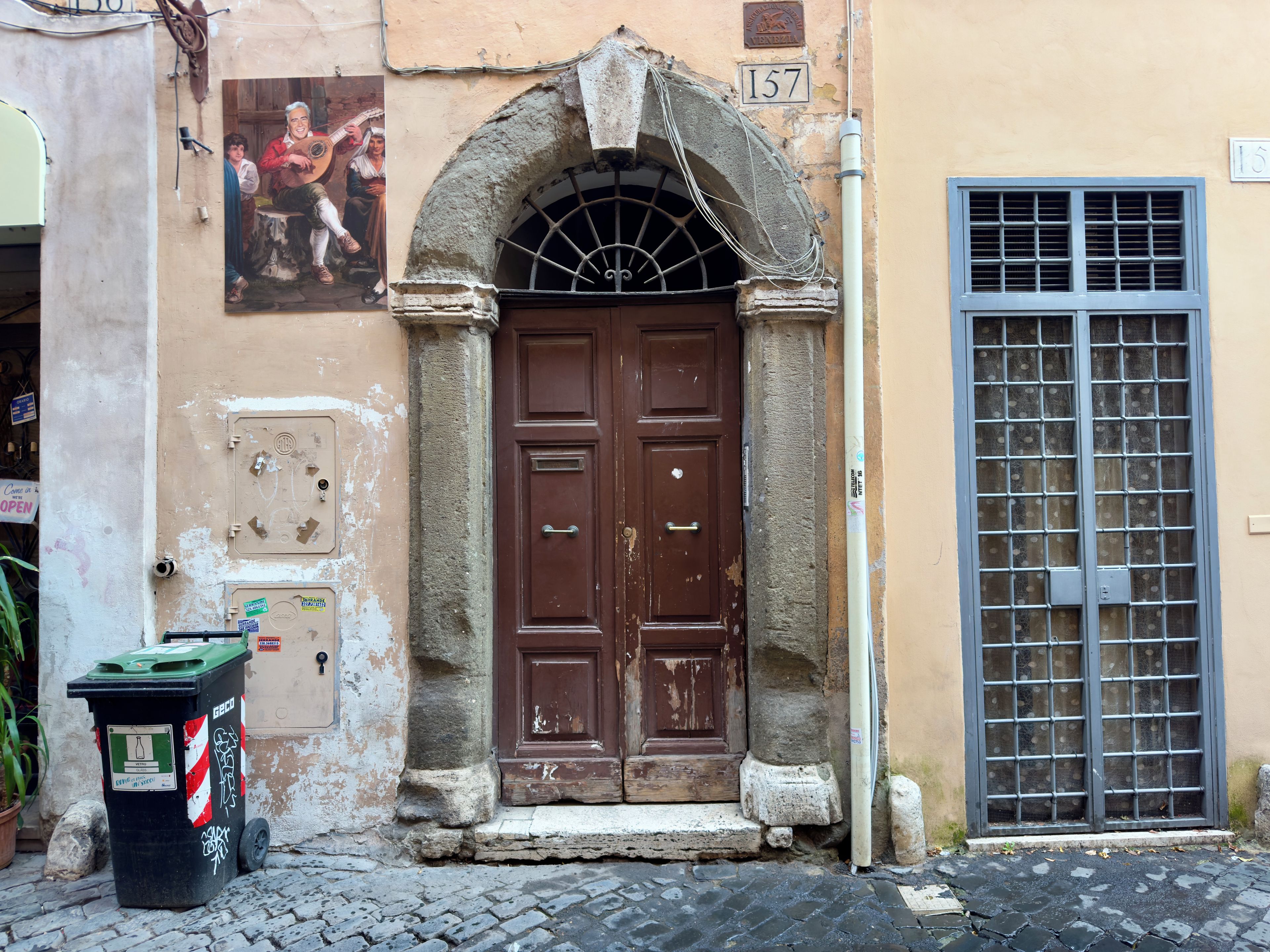 A brown door with a metal grille, a trash bin with recycling labels, and a window with a metal grid.