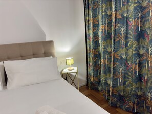 A bed with white linens, a headboard, a bedside table with a lamp, and a curtain with a tropical leaf pattern. - Vrbo Property (Roma)