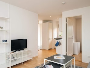 Ein modernes Wohnzimmer mit einem Flachbildfernseher, weißen Regalen, einem Couchtisch und einer Küche im Hintergrund.