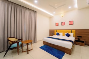 A hotel room with a bed, a chair, a small table, a ceiling fan, and wall-mounted lights. - Shri Kalyan Hotel And Restaurant Khatu (Khatoo)