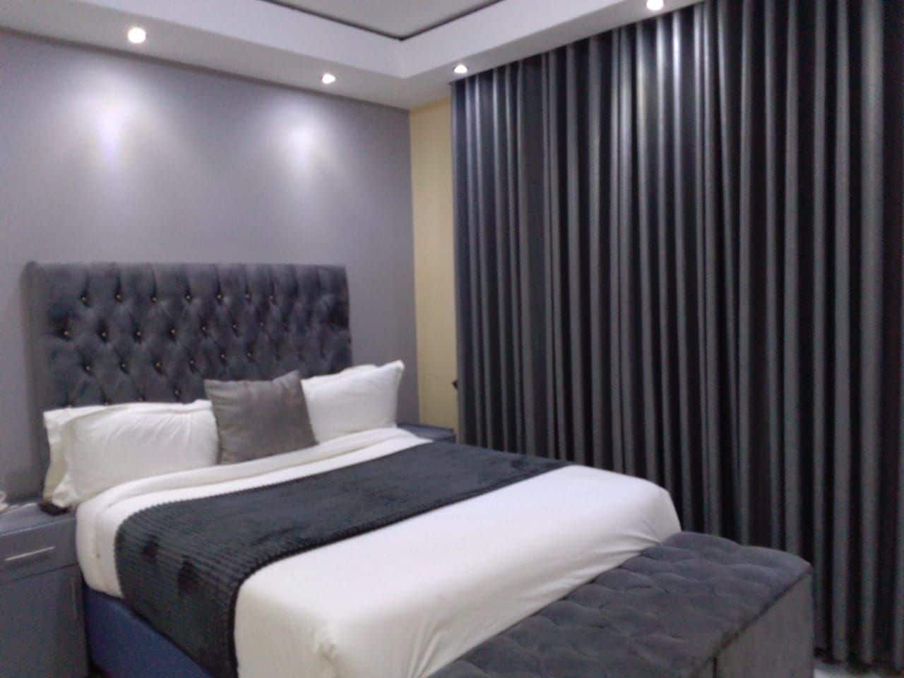 A modern bedroom with a tufted headboard, a large bed with white and grey bedding, and a dark curtain on the window.