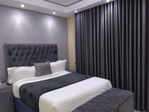 A modern bedroom with a tufted headboard, a large bed with white and grey bedding, and a dark curtain on the window. - Mazikana guest lodge, nestled high in the mountain, we offer best accommodation (Bindura)
