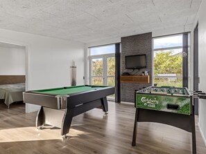 Ein moderner Raum mit Billardtisch, Kicker, Fernseher und einem Fenster mit Blick auf Grünflächen.