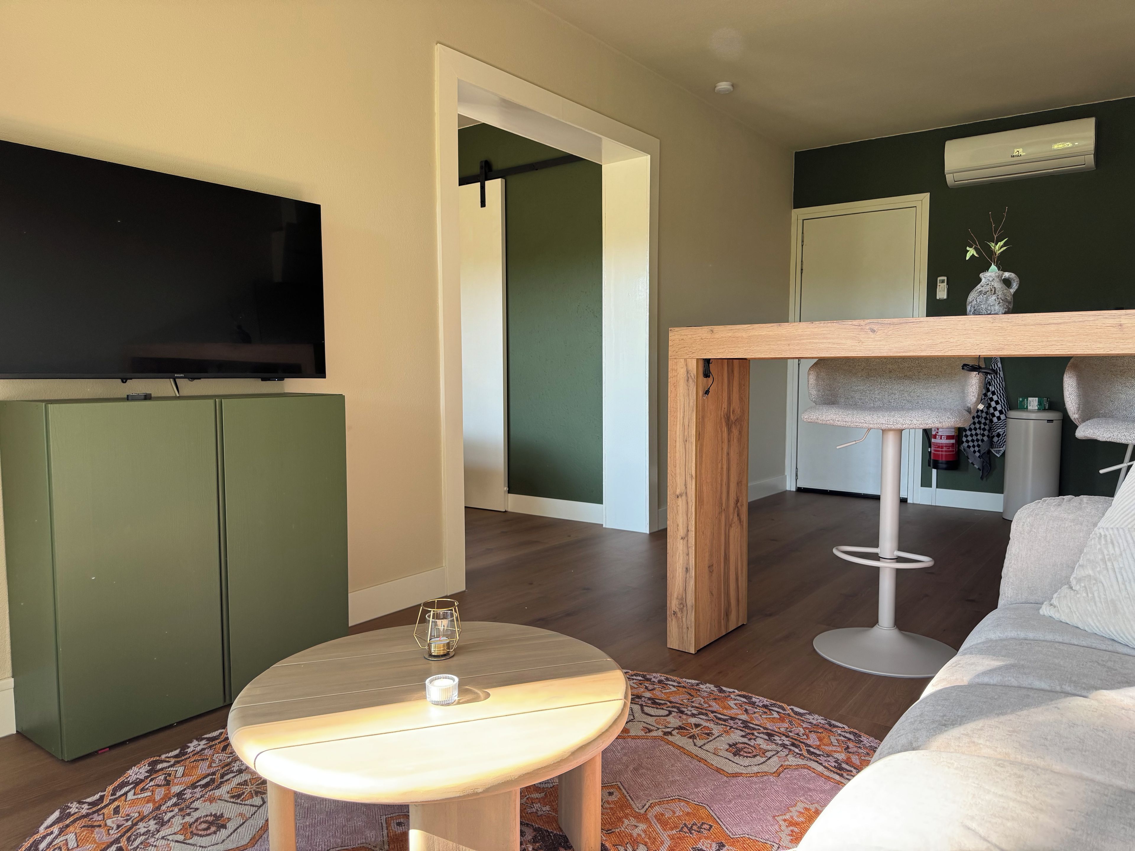 Ett modernt vardagsrum med en platt-TV, ett runt bord med en lampa, en soffa och en mönstrad matta.