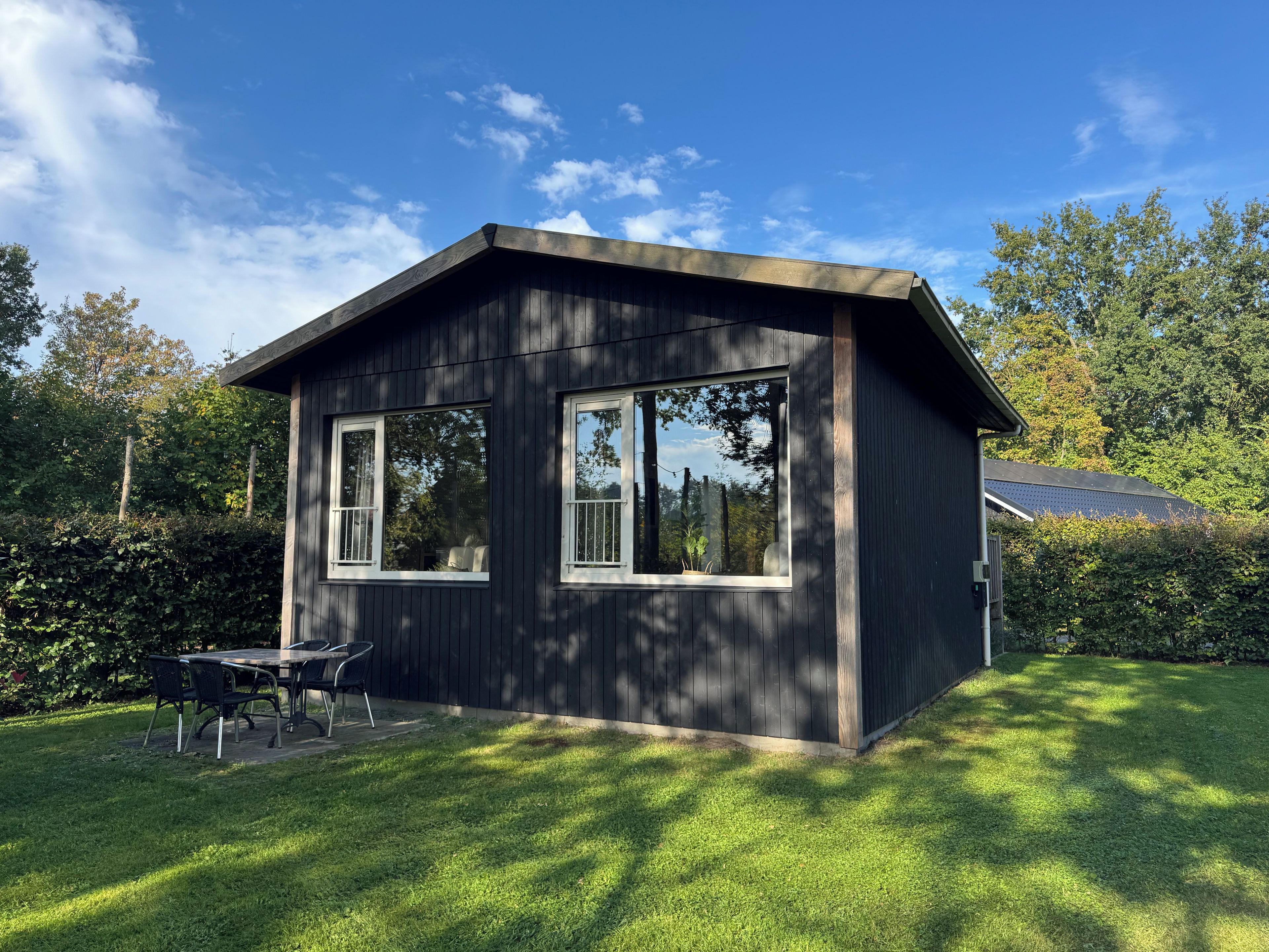 A small, dark wooden cabin with two windows and a patio area with a table and chairs.