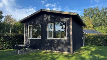 A small, dark wooden cabin with two windows and a patio area with a table and chairs.