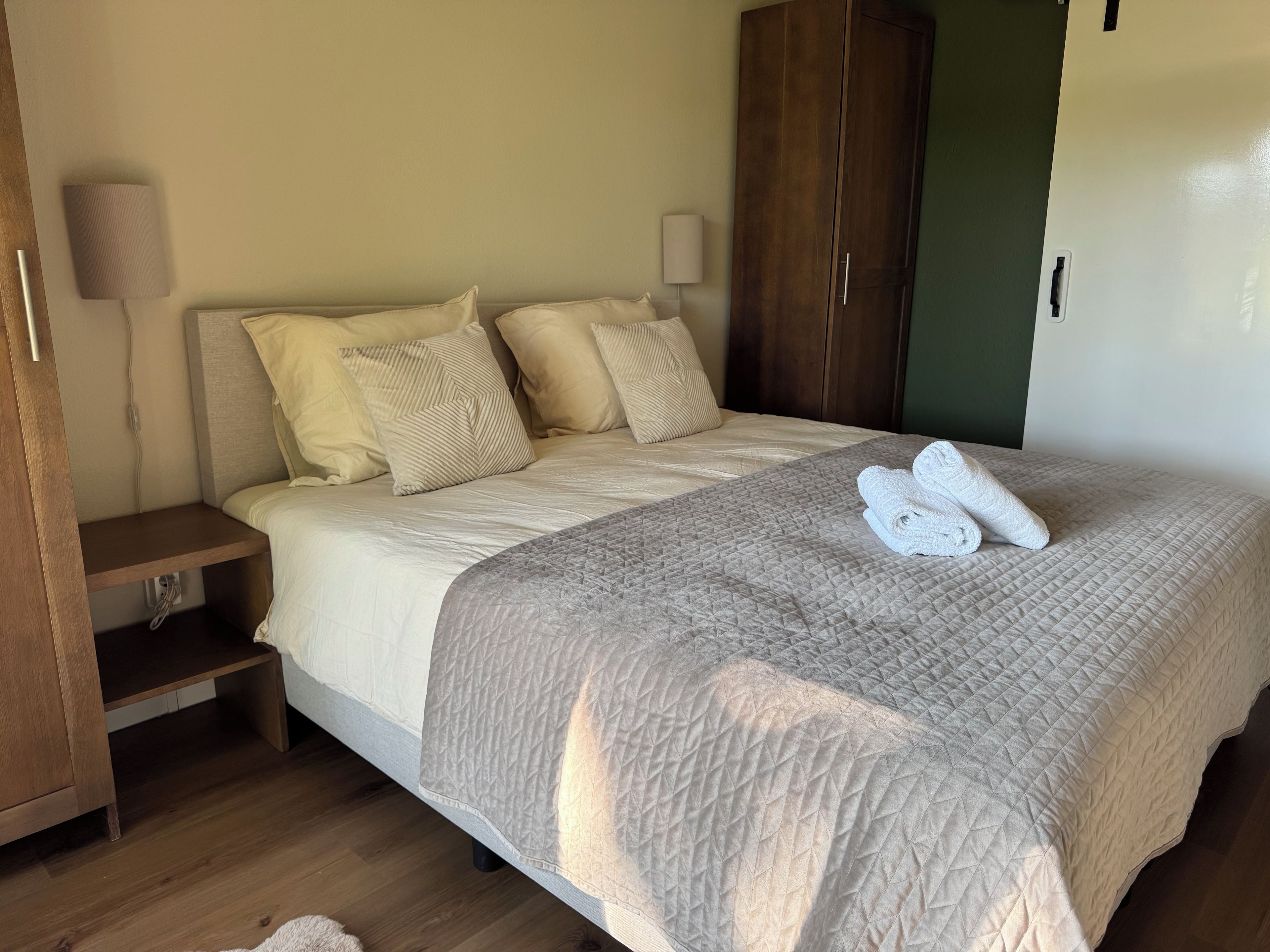 Un lit bien fait, recouvert d’une couverture matelassée, une armoire en bois et une lampe de chevet.