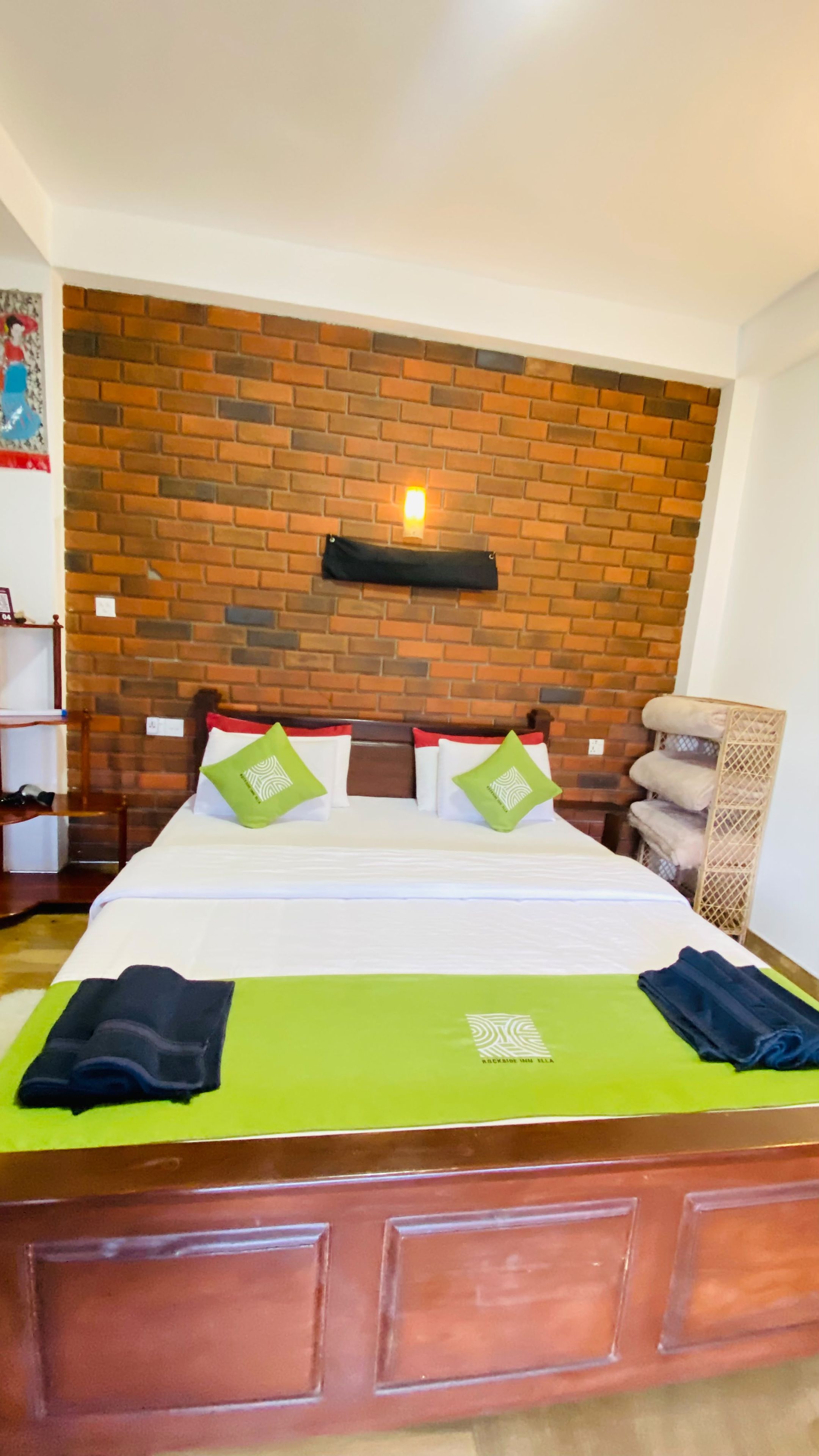 Una habitación de hotel con cabecera estampada con diseño de ladrillos, una cama bien hecha con ropa de cama verde y blanca, y una mesita de noche de madera con una lámpara.