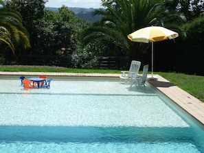 Una piscina con sillones de descanso y una mesa, rodeada de follaje y una valla.