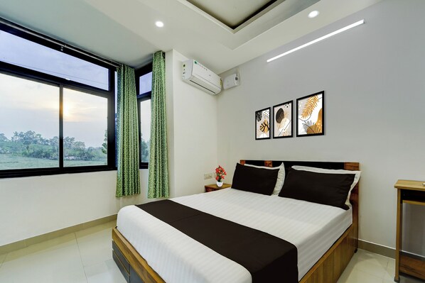 A hotel room with a large bed, a window with curtains, a wall-mounted air conditioner, and framed artwork.