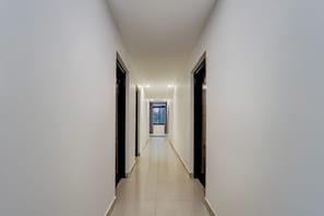 A long, narrow hallway with a tiled floor and white walls, leading to a room with a window.