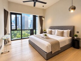 A modern bedroom with a large bed, a standing fan, a small table with a plant, and a view of the city through large windows.