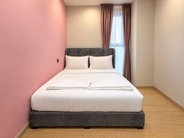A neatly made bed in a room with pink walls, a grey headboard, and a window with curtains.