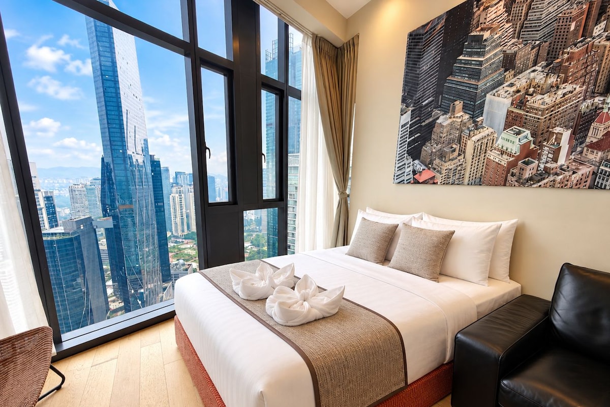 A hotel room with a large bed, a city view, a black leather chair, and a painting on the wall.
