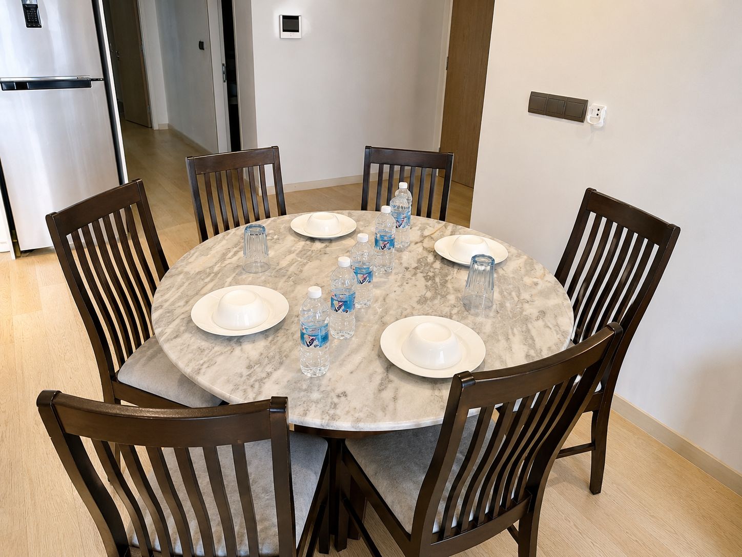 A round marble dining table with six chairs, surrounded by white plates and clear water bottles.