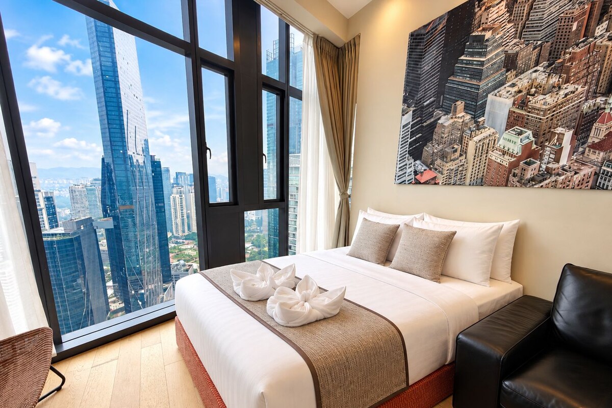 A hotel room with a large bed, a city view, a black leather chair, and a painting on the wall.