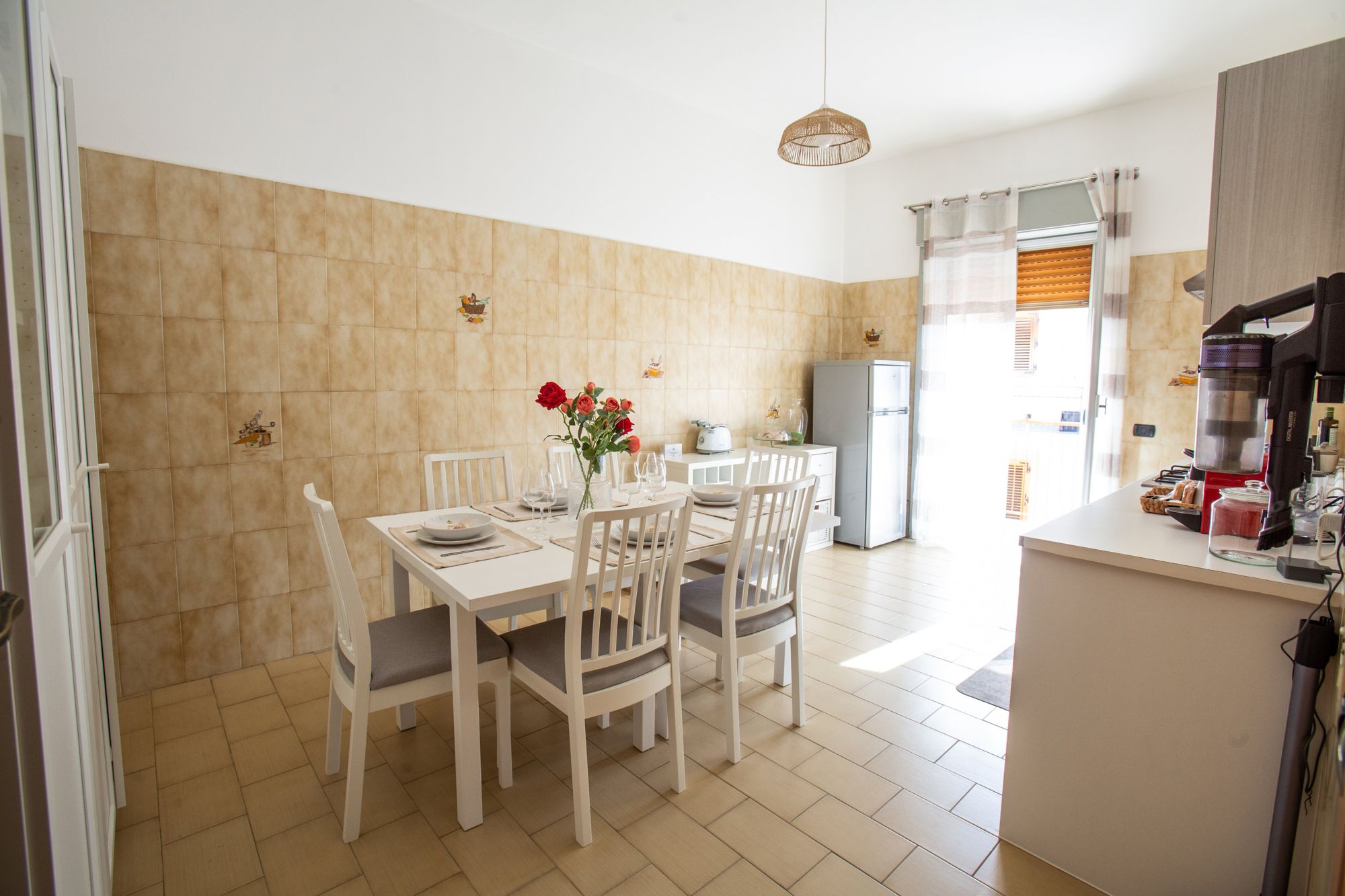 Un coin repas avec une table dressée pour quatre personnes, des chaises blanches, un vase avec des fleurs rouges et, au fond, une cuisine équipée d’un réfrigérateur et d’une machine à café.