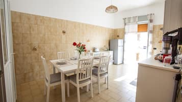 Un coin repas avec une table dressée pour quatre personnes, des chaises blanches, un vase avec des fleurs rouges et, au fond, une cuisine équipée d’un réfrigérateur et d’une machine à café.