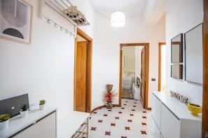 Un couloir avec un mur blanc, une porte en bois, une console blanche sur laquelle est posée une pancarte indiquant « BIENVENUE », et un sol à motifs.