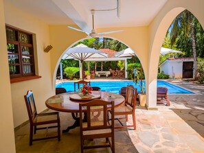 A poolside dining area with a table and chairs, a ceiling fan, and a view of the pool and surrounding greenery. - Spacious 4-bedroom paradise villa in Diani Beach with jacuzzi and private pool (Diani Beach)