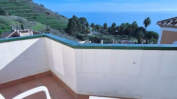 A terrace with two white plastic chairs and a small table, overlooking a coastal landscape with hills and palm trees.