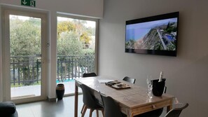 A room with a large window, a table with chairs, a television on the wall, and a bottle of champagne. - Il Giardino dell'Etna (Mascalucia)