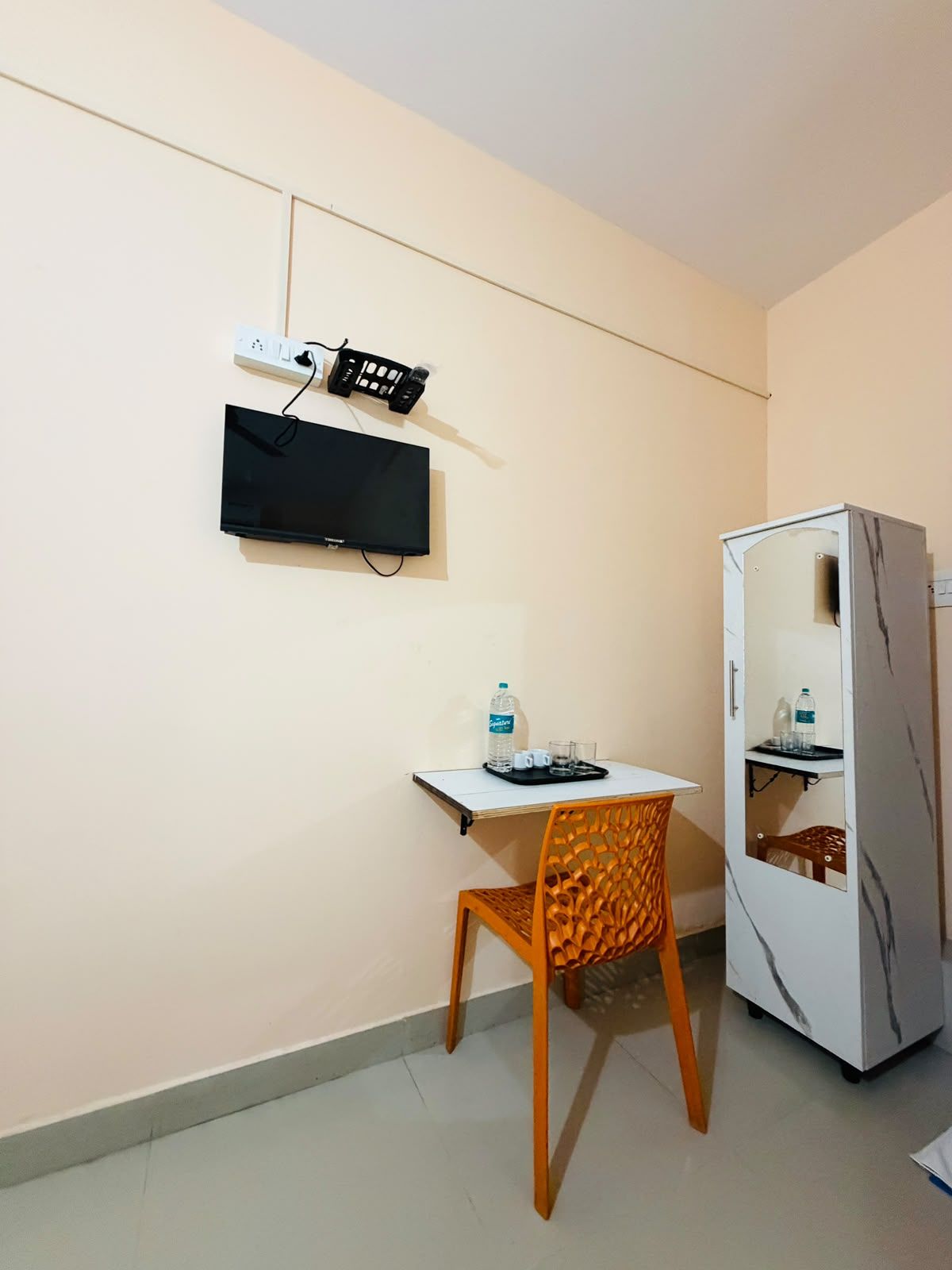 A small room with a white table, a chair, a television mounted on the wall, and a cabinet with shelves.